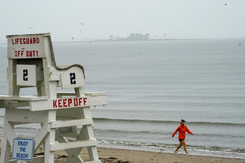 A woman in a red jacket and cap walks along the beach in the rain next to a white lifeguard chair.