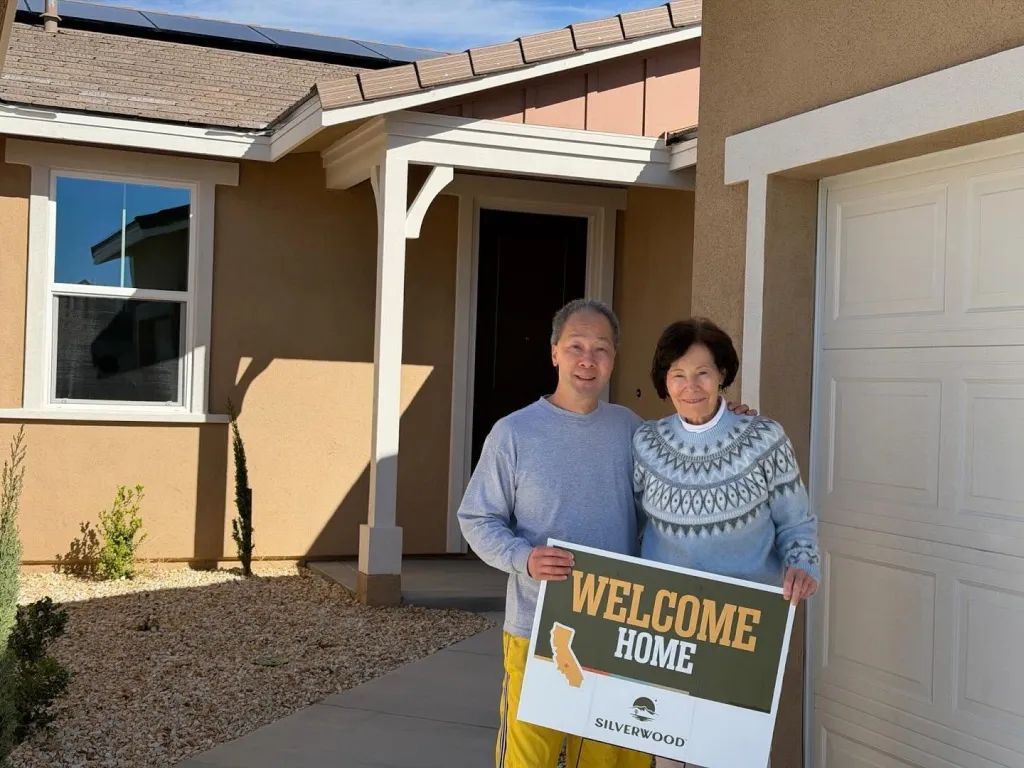 A smiling couple holding a "Welcome Home" sign in front of a new house.