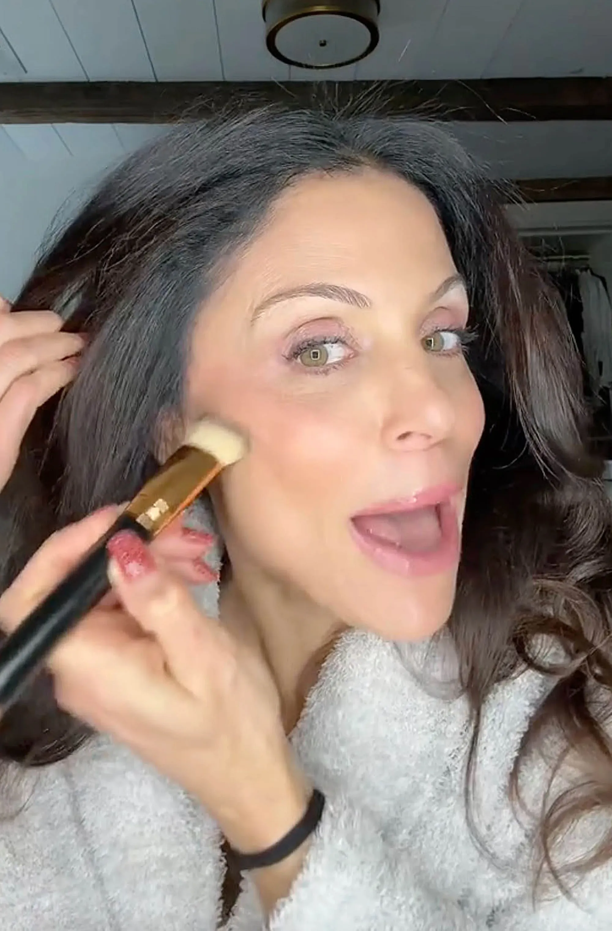 A woman with dark hair and a fluffy robe applies bronzer to her cheek with a makeup brush.