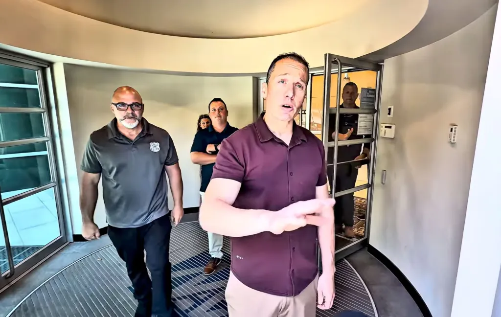 A group of men in casual clothing, one with a "PBA" emblem, confronting a man holding a camera.