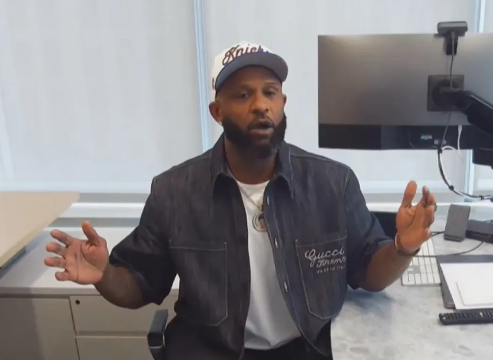 A man in a white cap, dark denim jacket, and white T-shirt sitting at a desk with a computer monitor.