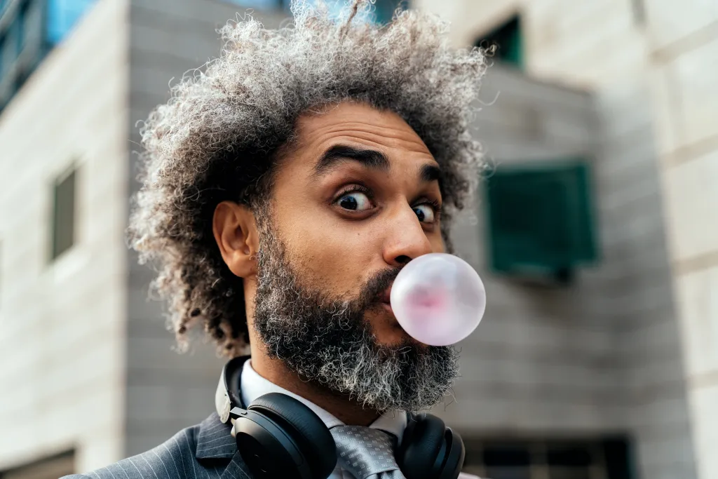 A man in a suit and tie with headphones around his neck blows a pink bubble gum bubble.