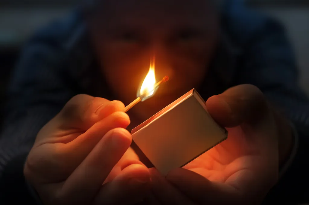 A person holds a burning match in one hand and a matchbox in the other, both hands lit by the flame in the dark.