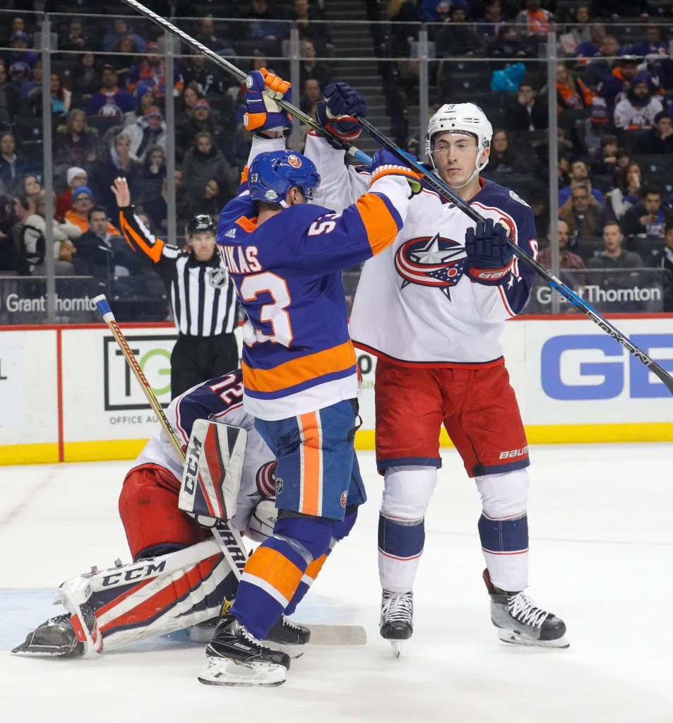 Hockey players Casey Cizikas #53 of the New York Islanders and Zach Werenski #8 of the Columbus Blue Jackets in a scuffle with a referee raising his hand in the background.