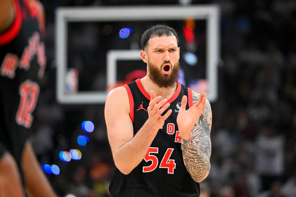 A man in a black and red basketball jersey with "54" on it claps his hands, looking intense, with a blurred basketball court in the background.