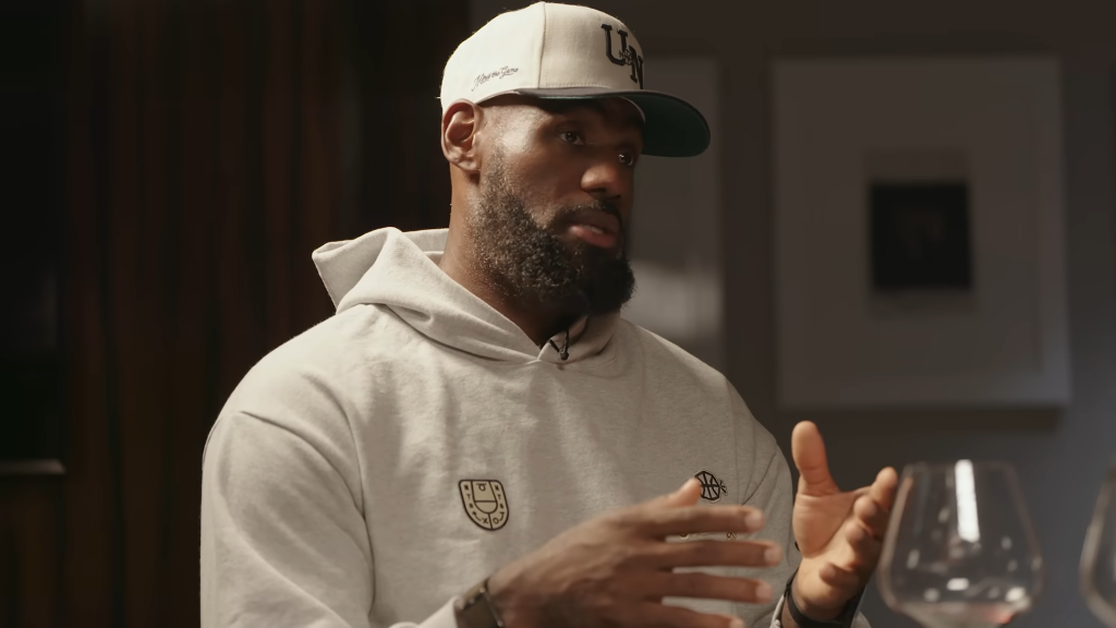LeBron James speaking while wearing a grey hoodie and a white and green baseball cap.