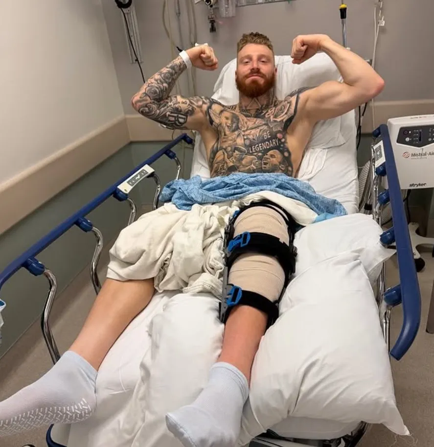 Maxx Crosby flexing his biceps in a hospital bed with a knee brace on his right leg.