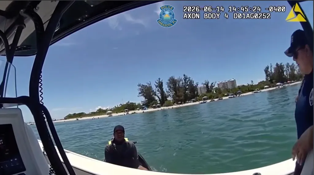Bodycam footage from a police boat on a sunny day shows an officer and a man in a life vest on a jet ski, with a beach and buildings in the background.