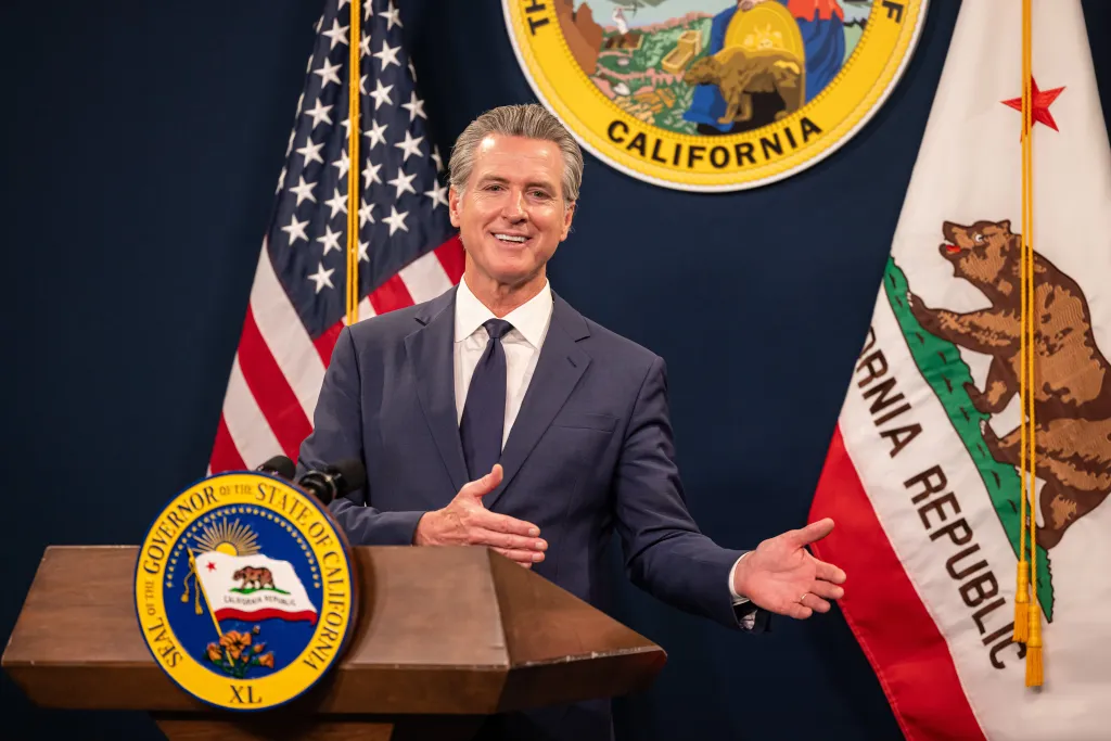 California Governor Gavin Newsom at a podium with US and California flags behind him.