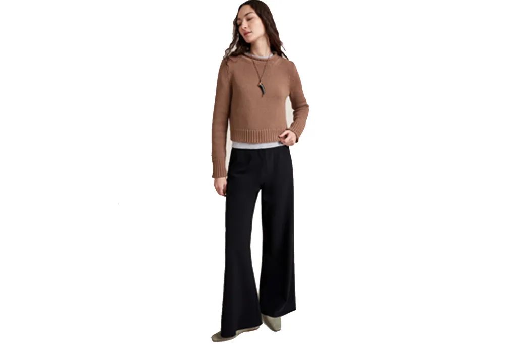 A woman in a cropped brown sweater, a gray top, black flared pants, and light green flats stands looking down to her left.