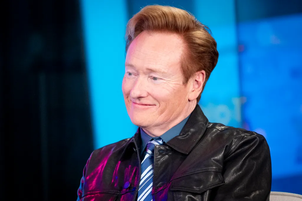 Conan O'Brien smiling while wearing a black leather jacket and blue striped tie.