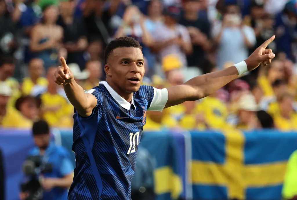 Kylian Mbappé in a blue and white uniform celebrates a goal, pointing to the sky with both hands.