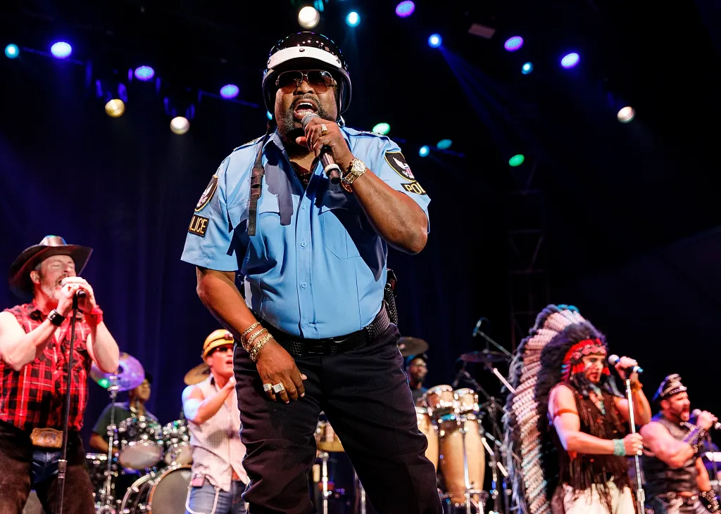Victor Willis of Village People performs on stage in a police uniform, with other band members visible.