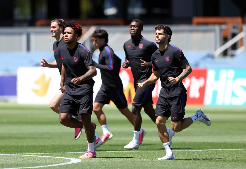 Five soccer players, including Weston McKennie and Christian Pulisic, run in a line on a sunny green field during a training session.